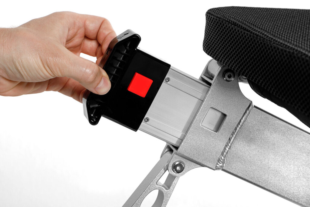 Porto Mobility Ranger Discovery folding power wheelchair, hand holding a pack with red button at the end of the frame tube below the seat