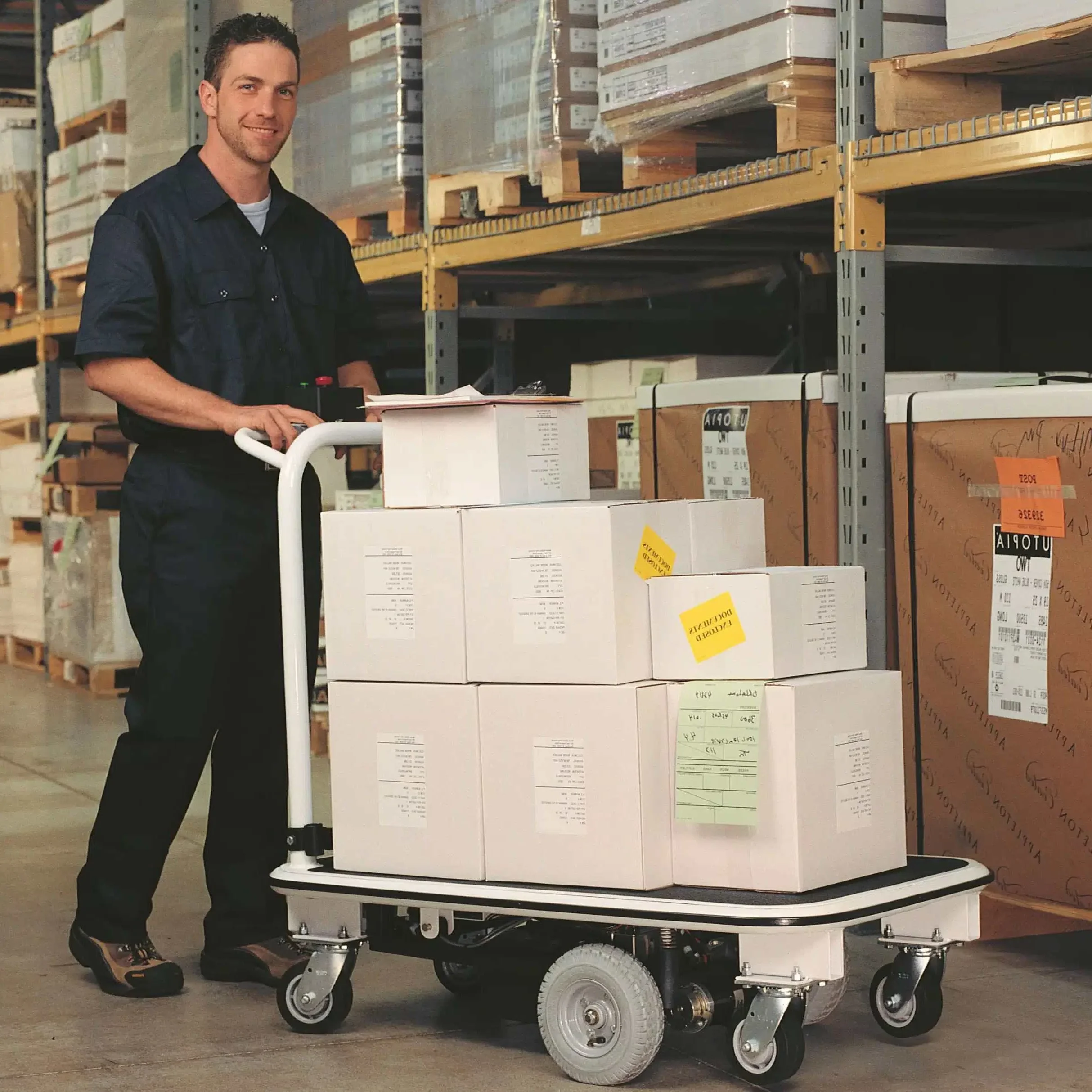Man in work uniform using EK Tech Pony Express 1031 motorized platform cart loaded with cardboard boxes in a warehouse aisle